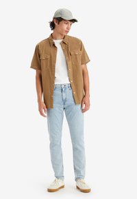 Brown short-sleeve shirt with two chest pockets, white t-shirt underneath, light blue jeans, and cream sneakers with a tan sole and cap.