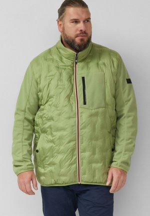 Mężczyzna z brodą w jasнозielonej pikowanej kurtce z suwakiem z przodu, kieszenią na piersi i granatowych spodniach, stojący na jednolitym tle.