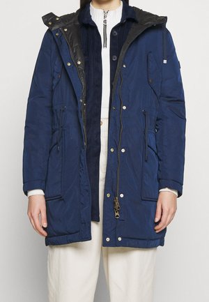 Femme portant un manteau bleu marine superposé avec fermeture éclair et boutons, taille resserrée par un cordon, sur un pull en maille blanche côtelée et un pantalon crème.