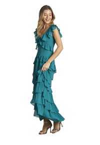 Abito maxi in teal, con volant, in tessuto a strati, scollatura a V profonda e maniche corte. Indossato con sandali con cinturini, che evidenziano movimento e consistenza.