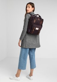 Backpack in dark purple fabric with brown leather accents, featuring two top handles and adjustable shoulder straps. Rectangular shape.