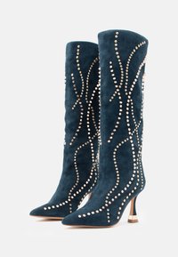 Knee-high, tumši tirkīza zamšādas zābaki ar smailu purngalu, rotāti ar zelta knaģiem viļņu veidā un smalku papēdi.