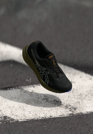 Chaussure de sport noire avec une tige respirante, semelle en caoutchouc et accents surélevés. Présente un design épuré avec une surface texturée et des détails réfléchissants.