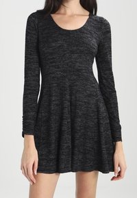 Donna che indossa un vestito in maglia grigio scuro a maniche lunghe, con scollatura rotonda e gonna svasata, in piedi davanti a uno sfondo semplice.