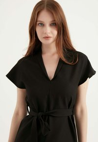 Schwarzes Kurzarmkleid mit V-Ausschnitt und einem gebundenen Stoffgürtel, aus glattem Material mit lässiger Passform und subtiler Textur.