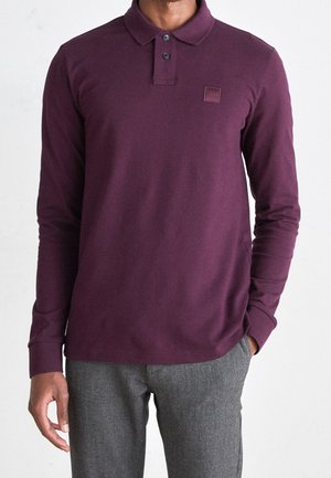 Mann trägt ein dunkelviolettes Polohemd mit langen Ärmeln und graue Hose, eine Hand in der Tasche, vor einfachem hellem Hintergrund.