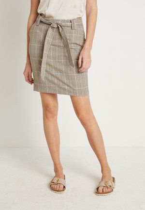 Frau trägt beige karierten Rock mit gebundenem Gürtel, weißes Oberteil und beige Sandalen mit großer goldener Schnalle, steht auf hellem Boden.