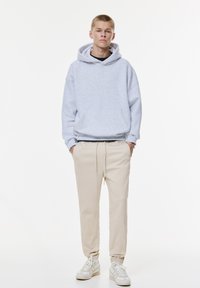 Felpa in pile grigio chiaro con tasca a marsupio, pantaloni jogger beige a taglio affusolato e scarpe da ginnastica bianche con dettagli grigi. Outfit semplice e informale.