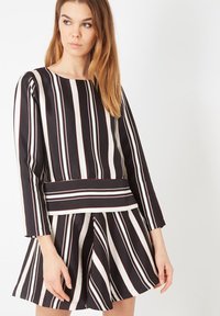 Mujer con blusa de manga larga de rayas verticales blancas y negras y falda acampanada a juego, de pie contra un fondo blanco.