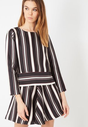 Mujer con blusa de manga larga de rayas verticales blancas y negras y falda acampanada a juego, de pie contra un fondo blanco.