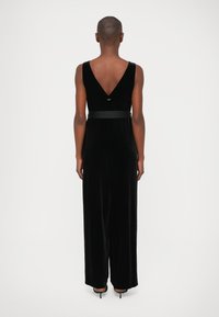 Mono de terciopelo negro con diseño de espalda en V, tirantes anchos y detalle de cintura ceñida. La tela tiene una textura suave y lisa.