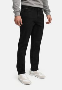 Mann trägt schwarze Jeans und weiße Turnschuhe, steht mit entspannten Händen an den Seiten vor einem einfarbigen Hintergrund.