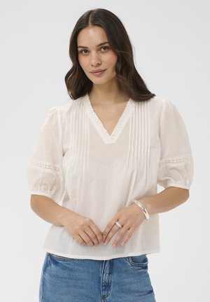 Femme aux longs cheveux foncés portant un chemisier blanc à col en V avec des manches bouffantes et un jean bleu, mains jointes montrant un bracelet et une bague.