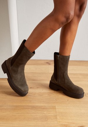 Bottines en daim vert olive avec une semelle noire épaisse, dotées de panneaux élastiques sur les côtés et d'une fermeture éclair pour un enfilage facile.