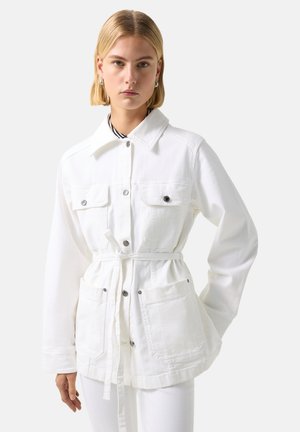 Junge Frau mit kurzen blonden Haaren, die eine weiße geknöpfte Jacke mit Gürtel und mehreren Fronttaschen trägt, steht vor einem schlichten Hintergrund.