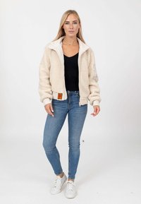 Giacca in pile soffice color crema con colletto alto, chiusura con zip e orlo a costine, abbinata a jeans skinny blu e sneakers bianche.