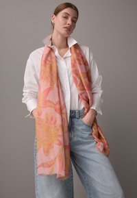 Vaaleanpunainen kukkahuivi, jossa on suuret keltaiset ja vaaleanpunaiset kukkakuvioinnit, valmistettu kevyestä kankaasta, puettu mallin kaulaan ja yhdistetty valkoiseen paitaan.