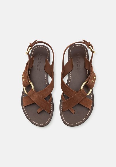 Brown suede sandals with crossed straps and a gold circular accent. Features a dark brown sole and white stitching. Adjustable ankle strap.