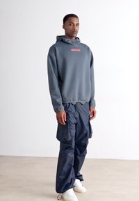 Un uomo che indossa una felpa grigia scura e pantaloni cargo neri con sneakers bianche, in piedi contro un muro bianco semplice.