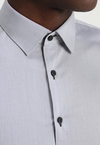 Vaaleanharmaa nappipaita, jossa on pehmeä kudos. Siinä on klassinen kaulus ja mustat napit, ja se esittelee yksinkertaisen mutta hienostuneen mallin.