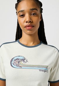 Camiseta color crema con ribetes azul oscuro que presenta un gráfico retro de surfista, diseño de olas y el logo de "Wrangler". Textura de algodón suave.