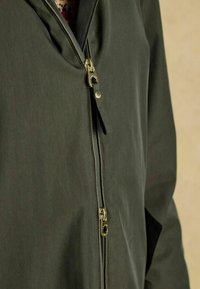 Olivengrøn jakke med lynlås lukning og guld hardware, et struktureret stof og et foret indre med et mønster, der er synligt ved kraven.