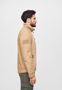 Tan fleece jacket with a high collar, front zip, and side pockets. Features a textured pattern and a fabric patch on the left sleeve.