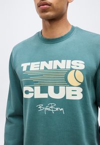 Άνδρας που φοράει τιρκουάζ φούτερ με το κείμενο "TENNIS CLUB" και κίτρινη γραφική απεικόνιση μπάλας τένις στο μπροστινό μέρος, με υπογραφή κάτω από το κείμενο.