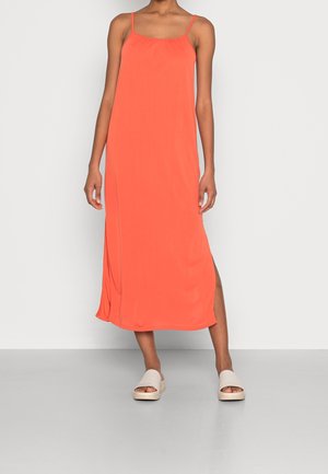 Femme portant une robe midi corail sans manches avec de fines bretelles et une fente latérale, associée à des sandales blanches à plateforme.