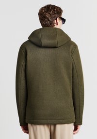 Mann in einer olivgrünen, strukturierten Kapuzenjacke und beigen Hosen, von hinten vor einem schlichten Hintergrund betrachtet.