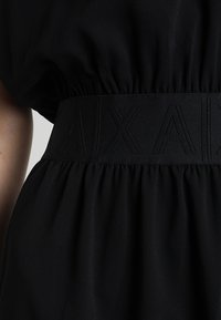 Gros plan sur une robe noire texturée avec une ceinture élastique présentant des lettres en relief subtilement embossées "A|X|A".