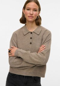 Brūns adīts džemperis ar apkakli, kuram ir trīs pogas, ribota tekstūra un garas piedurknes. Valkāts ar tumšiem džinsiem un melnu gredzenu.