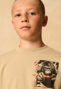 Bézs pamut póló, gorillát ábrázoló grafikával, amely fejhallgatót visel és játékkontrollert tart a kezében. Kerek nyakkivágás ribbelt textúrával.