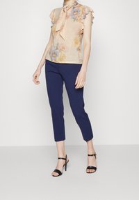 Kvinna iklädd en blommig ärmlös blus med en rosett, marinblå capribyxor och svarta högklackade sandaler står mot en enkel bakgrund.