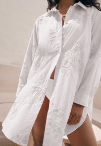 Chemise blanche brodée avec une coupe décontractée, ornée de motifs floraux, munie d'un col à boutons et de manches longues. Tissu léger et texturé.