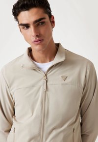 Beige zip-up jakke med en stående krave, to forlommer og et lille trekantet logo. Lavet af glat, letvægtsstof.