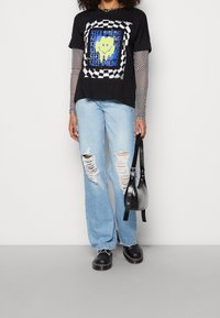T-shirt nera con stampa grafica, sovrapposta a una maglietta a maniche lunghe in rete, abbinata a jeans strappati di un blu chiaro e scarpe nere. Borsa nera piccola.