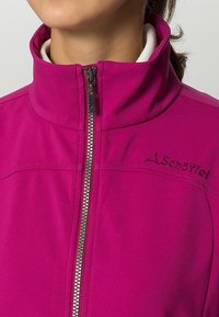 Mujer vistiendo una chaqueta softshell magenta con cremallera, cuello alto y el logo de Schoffel en el pecho.