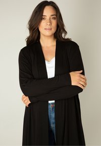 Vrouw met lang donker haar, gekleed in een zwarte openvallende cardigan over een witte top en een blauwe spijkerbroek, staand met gekruiste armen tegen een effen achtergrond.