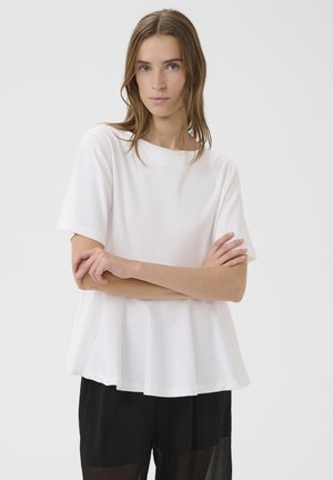 Mujer joven con cabello castaño claro, vestida con una blusa blanca de manga corta y suelta y pantalones negros, de pie con los brazos cruzados contra un fondo liso.