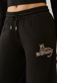 Melni sporta bikses ar elastīgu jostu un auklu, uz kurām ir spīdīga leoparda druka un "True Religion Brand Jeans" uzraksts uz augšstilba.