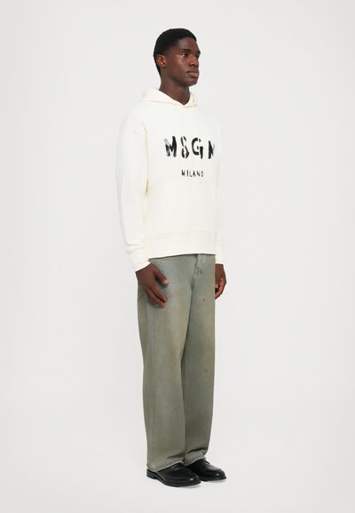 Giovane uomo che indossa un hoodie bianco MSGM Milano, jeans verdi sbiaditi e larghi, e scarpe di pelle nere, in piedi contro uno sfondo bianco semplice.