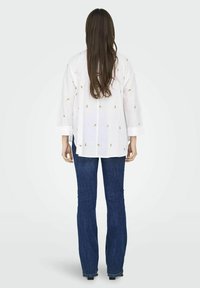 Camicia bianca ampia con ricami floreali gialli e verdi, abbinata a jeans a palazzo blu scuro. Tessuto morbido e design casual.