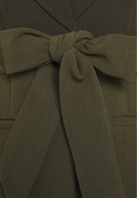 Olivgrünes Wickelkleid mit strukturierter Oberfläche, das mit einem geknoteten Gürtel an der Taille und zwei Vorderentaschen versehen ist.