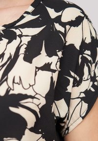 Schwarze und cremefarbene Bluse mit floralem Muster, glatter Textur, kurzen Ärmeln und rundem Ausschnitt, der mit kräftigen, abstrakten Blütenblattformen verziert ist.