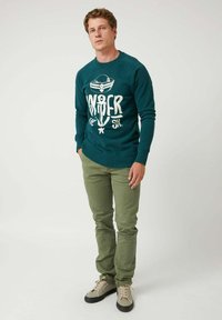 Hombre de pie vistiendo un sudadera azul marino con un ancla y el texto "Bajo el mar", pantalones verde oliva y zapatillas beige frente a un fondo blanco.