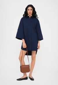 Abito camicia oversized blu navy con colletto a punte, chiusura frontale con bottoni, maniche corte e spacco laterale. Abbinato a una borsa a tracolla marrone.