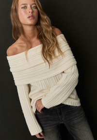 Oprijet pulover s smerami z raglani iz kremne barve z teksturirano tkanino in širokimi manšetami, kombiniran s temno sivimi jeans hlačami, z razbremenjenim krojem.