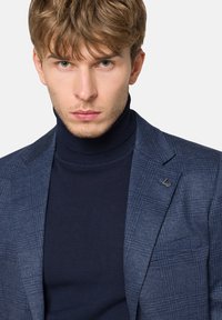 Granatowa marynarka w kratę z klapami, noszona na granatowym golfie. Posiada przypinkę z logo na klapie marynarki.