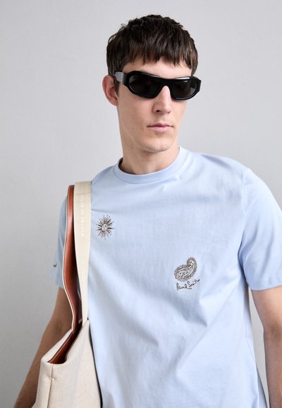 Ljusblå bomullströja med sol- och paisleymönster i brunt, bärs med svarta solglasögon och en beige tygväska med röd foder.